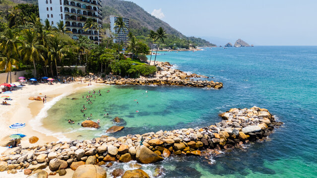 Aerial view of Playa Esmeralda with people swimming in turquoise water, rocky breakwaters, and beachfront resorts in Puerto Vallarta, Jalisco, Mexico.