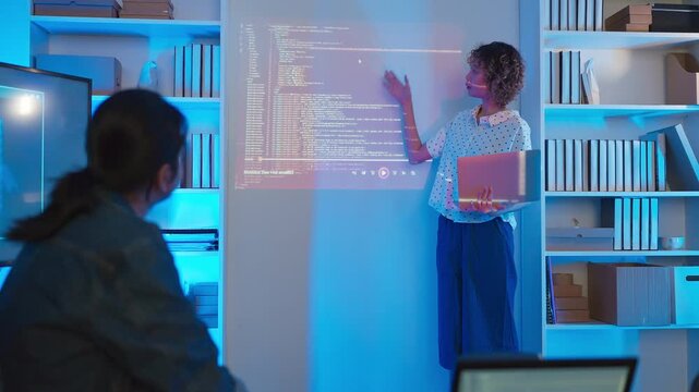 A professional East Asian female software engineer stands holding a laptop and gesturing toward lines of code projected on a wall display, presenting data scripts to her multiethnic colleagues during 