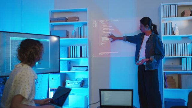 A professional male software developer stands presenting a data script and system infrastructure to his colleague during a late-night tech briefing, looking at camera while lines of code are projected