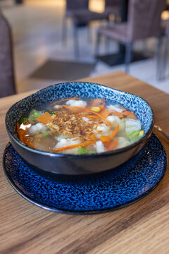 Warm soup with carrots, corn, and greens. Blue speckled bowl sits on matching plate. Wooden table and blurred chairs in background. Garnished with toasted croutons on top. Cozy, inviting meal setting