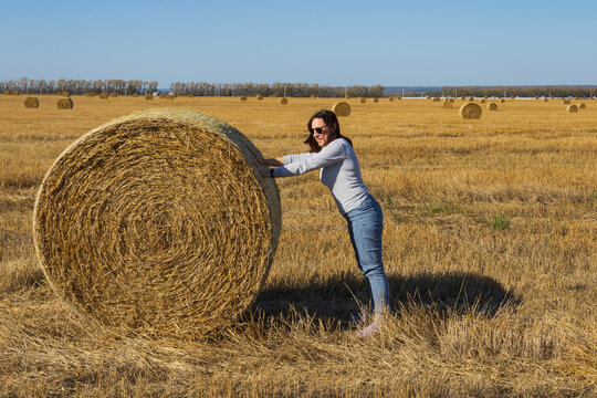 Young woman in casual clothes pushing a huge round straw hay bale in a golden agriculture field under a clear blue sky, harvesting crops at organic farm during sunny autumn day for eco tourism and rur