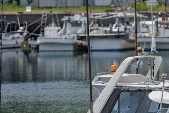 Atmospheric marina scene focusing on a white boat's cabin adorned with an orange beacon light, loudspeaker, and antennas, set against a bokeh background of other fishing boats moored in calm harbor.