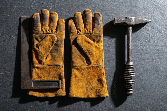 Top-down view of heavy weathered ochre leather welding gauntlets lying beside iron slag hammer and angle square tool on dark slate mineral composite worktop texture.