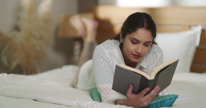Indian young adult gen z relax calm woman lying on cozy bed read new novel book story indoor room resting girl spend free time do self care enjoy day take break from job work home place