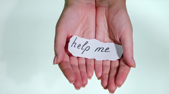 A desperate plea help me message, help me written on a piece of paper displayed in cupped hands of a young woman, white background, slow motion, close up.