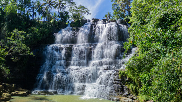 Aerial view of Cigangsa Waterfall with cascading water over tiered rock cliffs surrounded by lush tropical jungle and palm trees Sukabumi, West Java, Indonesia.