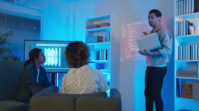 A professional male software engineer stands presenting and explaining lines of code projected on a wall display during a technical meeting, gesturing towards the data script in a dark room with drama