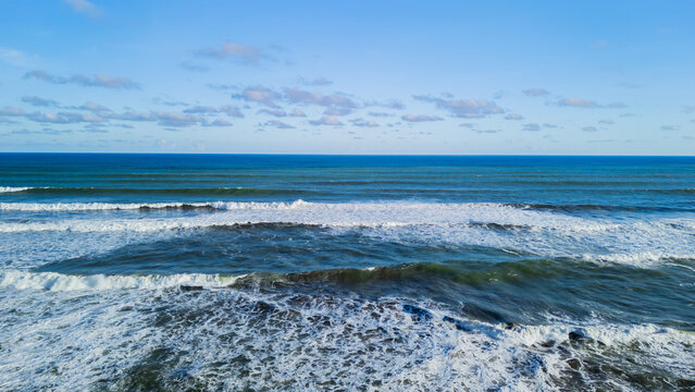 Aerial view of Ujung Genteng Beach with rolling ocean waves, white sea foam, and a clear blue sky with scattered clouds in Sukabumi, West Java, Indonesia.