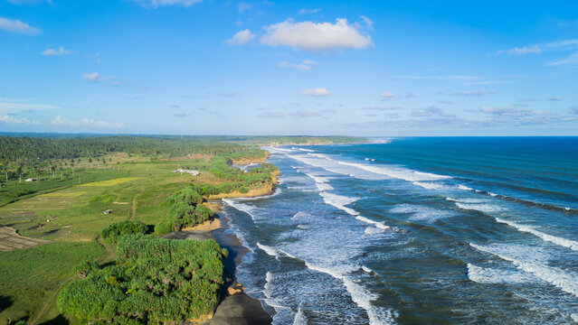 Aerial view of Ujung Genteng Beach with white waves crashing on the dark sand shore and lush green vegetation in Sukabumi, West Java, Indonesia.