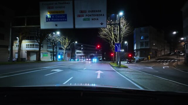 Driver's point of view driving at night in a city, approaching an intersection and stopping at a red traffic light with other vehicles waiting, illuminated by streetlights and buildings