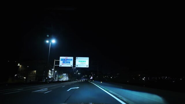Driver's perspective while navigating an empty motorway with headlights on, passing under glowing streetlights and directional signs on a dark evening, showing the quiet side of commuting