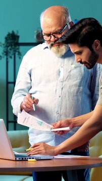 Indian young son and senior father filing online tax return together using laptop while standing at table in modern home, discussing financial reports, income statements yearly accounting documents