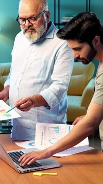 Indian young son and senior father filing online tax return together using laptop while standing at table in modern home, discussing financial reports, income statements yearly accounting documents
