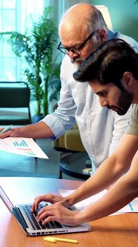Indian young son and senior father filing online tax return together using laptop while standing at table in modern home, discussing financial reports, income statements yearly accounting documents