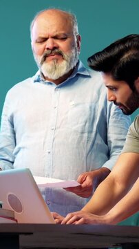Indian young son and senior father filing online tax return together using laptop while standing at table in modern home, discussing financial reports, income statements yearly accounting documents