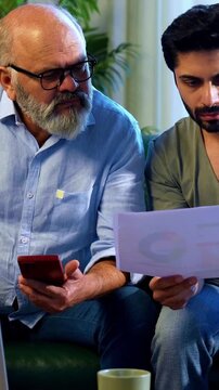 Indian retired father and young son working together on laptop and smartphone at home, reviewing financial report, share trading data and online business documents while discussing profit and loss