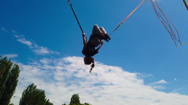 Low angle static shot captures a child harnessed into an elastic bungee system, performing forward and backward flips high in the air against a bright blue sky at a springtime park festival.