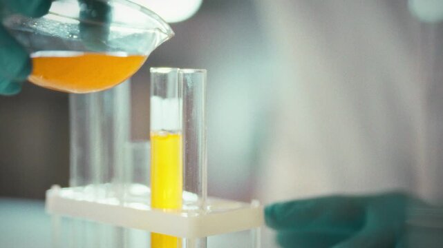 In a laboratory filled with gleaming equipment, a researcher in a white coat and protective gloves carefully pours liquid into a test tube, conducting a controlled experiment with precise technique