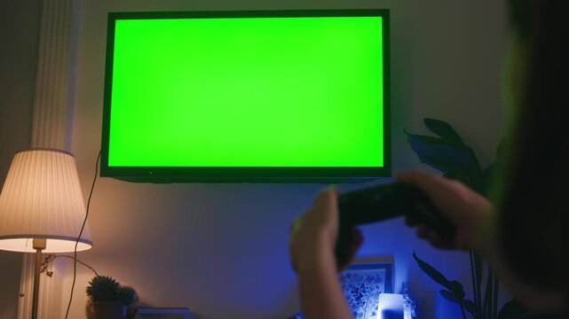 Girl sitting in the living room holding a game controller and playing video games, TV with a green screen. High quality 4k footage