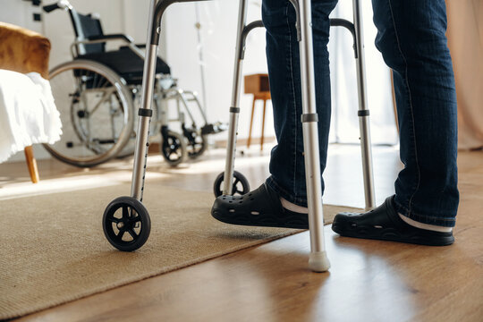 Individual using a walker for support stands on wooden floor in a well-lit room with a wheelchair visible in the background and a cozy chair nearby