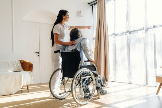 Female caregiver assists a man in a wheelchair, pointing towards sunlight streaming through large windows in a bright, modern living room setting