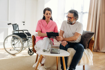 Fototapeta premium Female caregiver in pink scrubs assists elderly man with tablet in a cozy living room, wheelchair positioned nearby, sunlight streaming through large windows