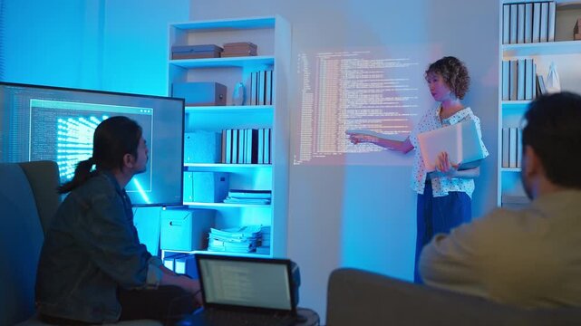 A professional East Asian female software engineer stands holding a laptop and gesturing toward lines of code projected on a wall display, presenting data scripts to her multiethnic colleagues during 