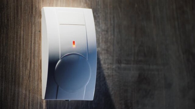 White plastic motion sensor on a brown wall, security alarm detecting movement, blinking red light indicates an alert.