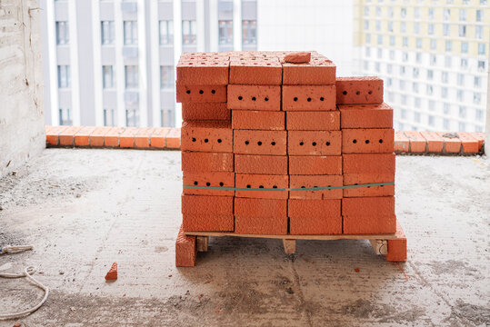 A stack of red perforated clay bricks prepared on a makeshift pallet on a concrete floor inside an unfinished high-rise building with an urban cityscape view.