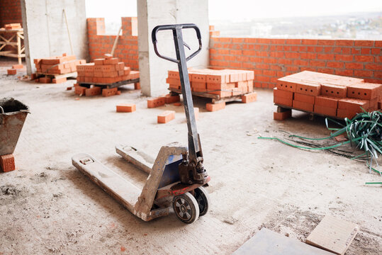 A dirty manual pallet jack stands on a concrete floor among stacked red clay bricks and a mortar tub inside an unfinished high-rise building construction site.