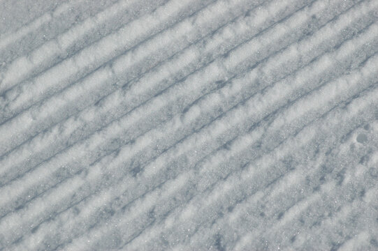 Full frame background texture of freshly groomed white winter snow showing a distinctive parallel corduroy pattern on a ski slope under sunlight