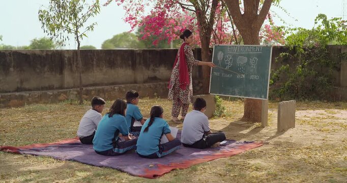 Indian young adult female tutor hold stick pointing words teach to girl boy under tree rural area place village group sitting on mat look board talk speak read do study learn open ground class field