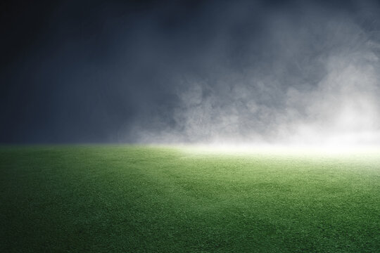 An empty green grass turf field illuminated by a bright spotlight under a dark, smoky, and misty night atmosphere, ideal for sports or event background