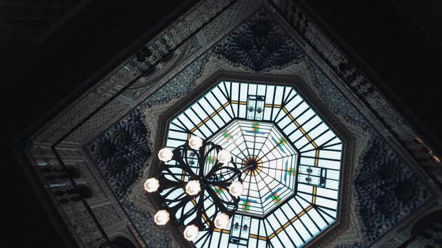Low angle view of an elegant ornate arabic patio ceiling featuring a stunning octagonal glass skylight dome and a beautiful vintage chandelier illuminating dark intricate architectural details and dee