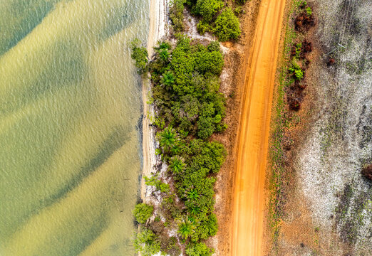Aerial view of a bright orange dirt road running alongside a tropical coastline with lush green trees and clear shallow water Monrovia, Liberia.