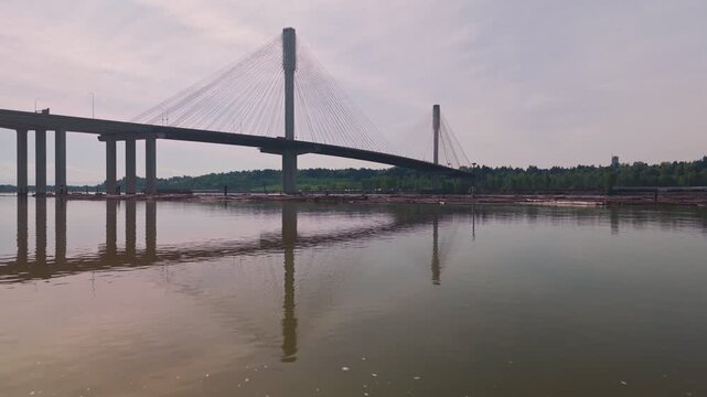 Cable stayed bridge towers dominate horizon while camera glides low over glassy river water reflecting massive architectural span with floating log booms near forested shoreline under hazy sky approac