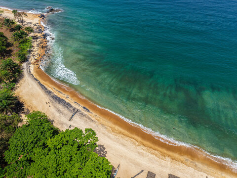 Aerial view of a tropical coastline with turquoise ocean waves washing onto a sandy beach lined with lush green trees in Monrovia, Liberia.