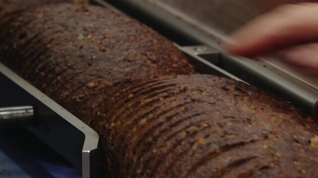 Close-up of a crusty dark bread loaf being sliced on bakery equipment, showing textured crust, grains, and commercial production detail.
