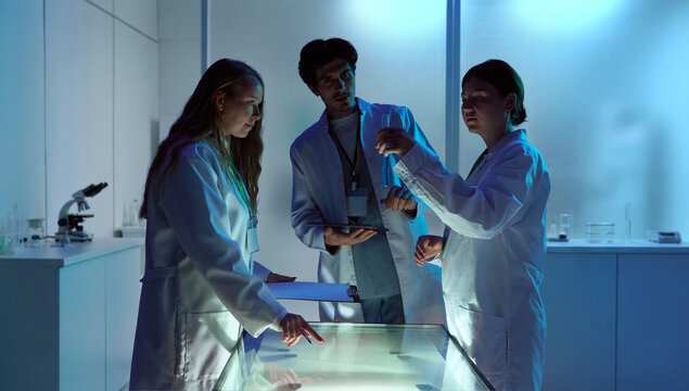 Laboratory team discussing chemical sample under blue research lighting. Concept of clinical testing, pharmaceutical research, lab teamwork, and biotech development.