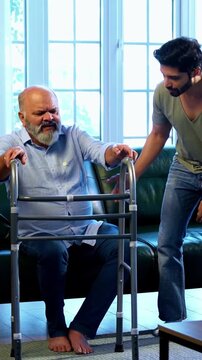 Indian young son helping and assisting aged father stand and walk with support of medical walker in living room at modern home, showing care, patience and responsibility toward elderly parent