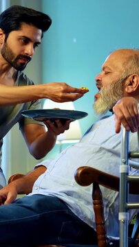 Indian young son feeding senior father chapati with sabzi from plate while he relaxes on chair, sharing heartfelt conversation and affection in modern home living room during homely mealtime