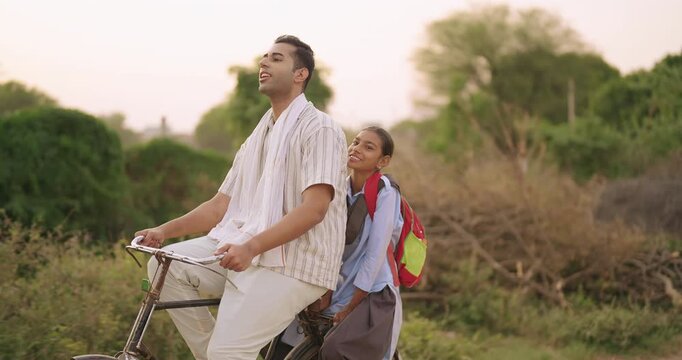 Indian happy young adult dad talk speak drop school to teen girl child rural area place road path way. Smiling female wear uniform with bag sitting on cycle back seat enjoy ride have fun joy desi life