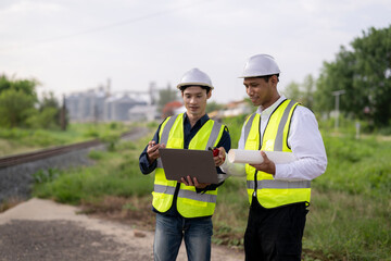 Obraz premium Two construction engineers in safety gear using tablet and clipboard outdoors near railway tracks