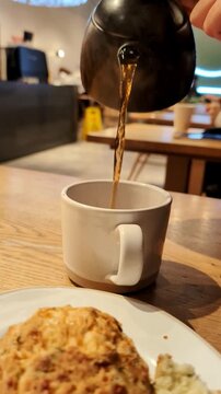 Person pouring hot tea from a traditional japanese teapot into a white ceramic cup during breakfast in a modern cafe with a scone on the side, showing a relaxing moment