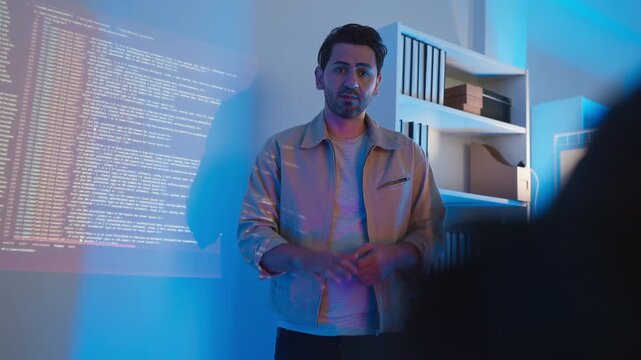 A professional male software engineer stands presenting and explaining lines of code projected on a wall display during a technical meeting, gesturing towards the data script in a dark room with drama