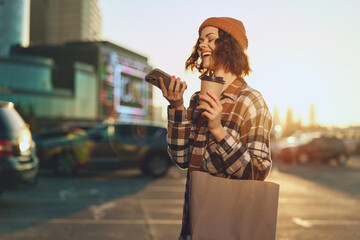 Woman holding coffee and phone with a bright smile on an urban street at golden hour sunlight,...