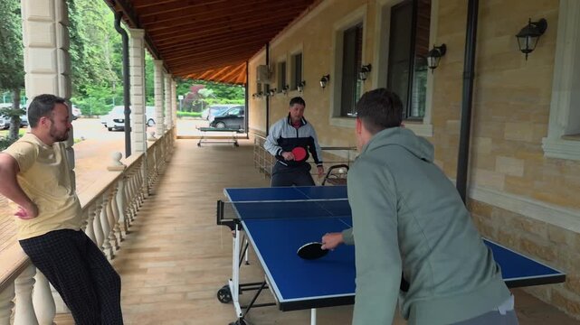 Adult son leans into playful ping pong rally with laughing father on covered veranda as friend watches by railing, handheld rear view beside blue table, stone wall, wooden roof and garden trees