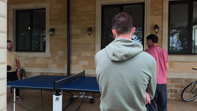 Authentic Men Playing Table Tennis on Covered Stone Terrace, Spectator Seen from Behind Watching Ping Pong Rally, Blue Outdoor Table, Side Tracking Handheld Shot in Soft Natural Daylight by Windows
