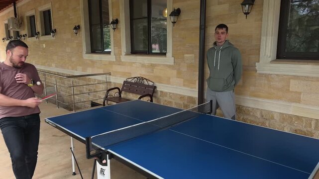 Beginner man awkwardly missing ping pong ball and learning to serve during casual table tennis game with family on covered house terrace by stone wall, windows and porch lights in lifestyle shot