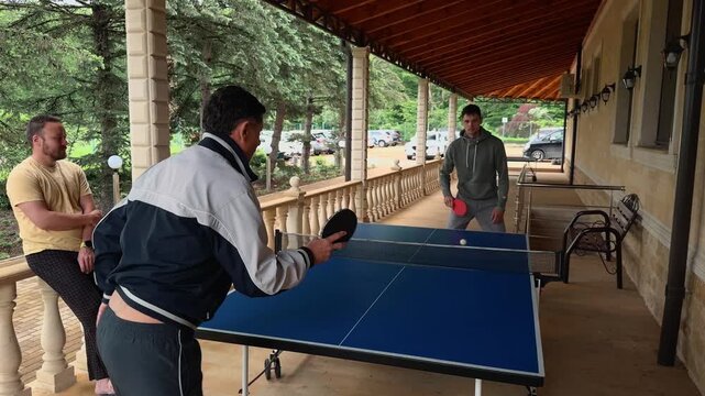 Intergenerational ping pong rally on covered veranda, older father serving while son waits and family friend watches, handheld view beside blue table, stone house wall, wooden roof, columns and trees
